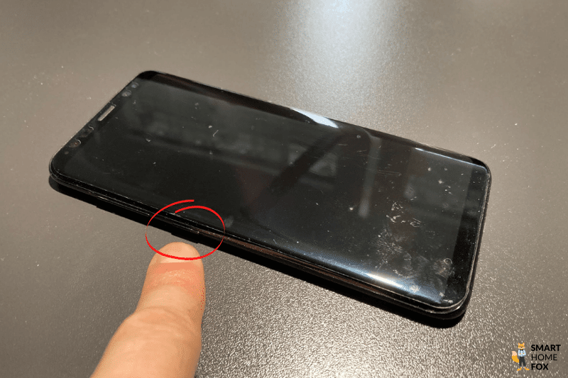Ein Samsung Handy ist von der Seite zu sehen, wobei der mit dem Zeigefinger auf dem Bixby Button gezeigt wird
